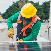 A worker in a yellow hard hat installs or inspects solar panels on a rooftop, wearing a green shirt, orange-trimmed vest, and gloves. Trees and a building are visible in the background.